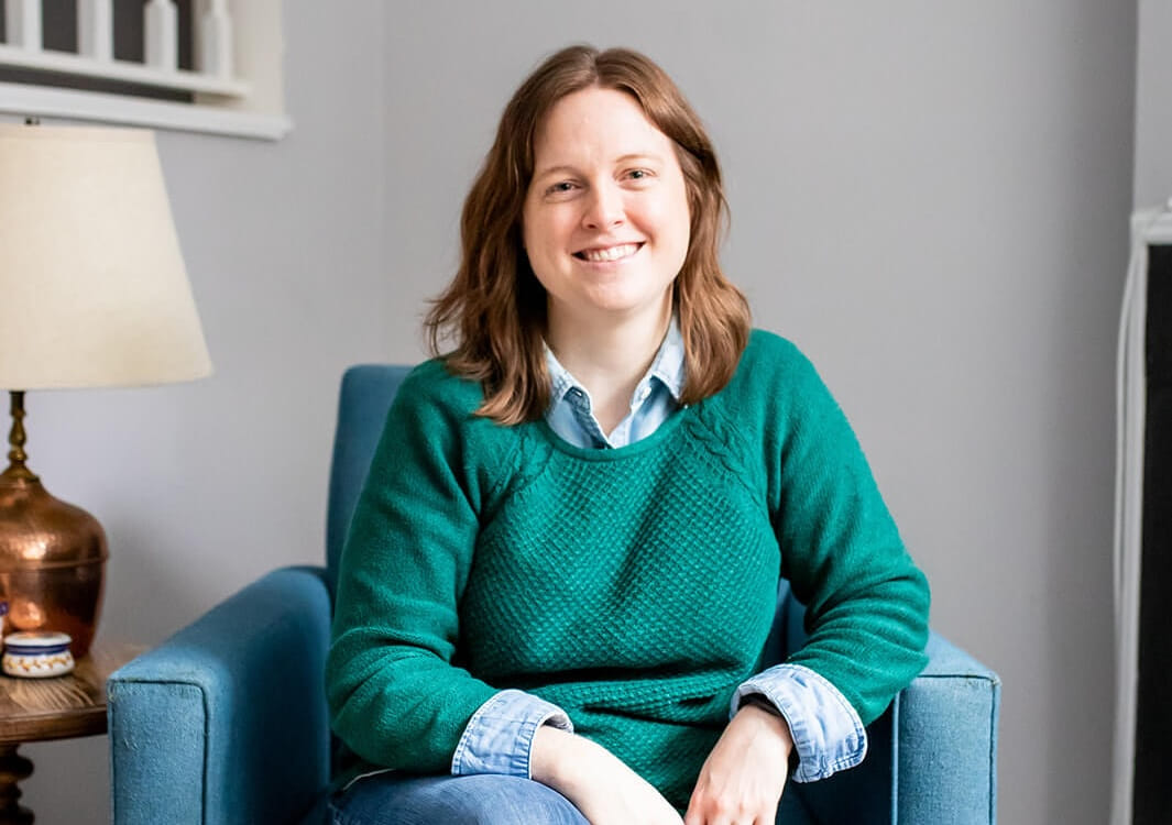 Emily Laing, LPC, a white woman in her mid to late 30s with shoulder-length auburn hair, smiles towards the camera. She wears a grey wool coat over a green sweater and denim collared shirt. She stands outside in front a tree and snow.