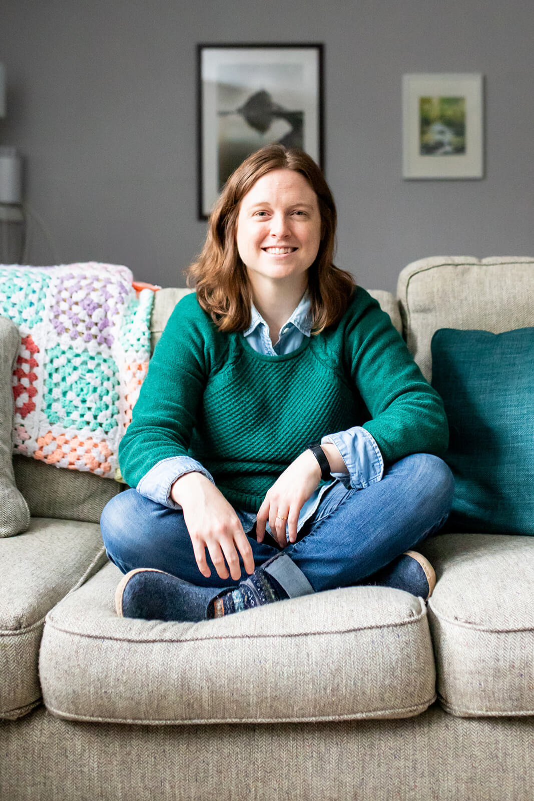 Emily Laing, LPC, a white woman in her mid to late 30s with shoulder-length auburn hair, smiles towards the camera. She wears a green sweater with a denim collared shirt underneath, jeans, and blue slippers. She sits cross-legged on a beige couch.