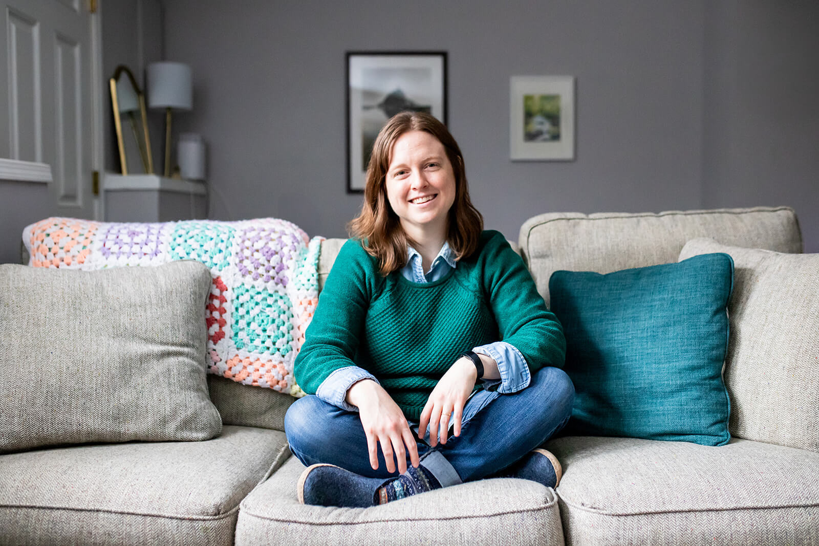 Emily Laing, LPC, a white woman in her mid to late 30s with shoulder-length auburn hair, smiles towards the camera. She wears a green sweater with a denim collared shirt underneath, jeans, and blue slippers. She sits cross-legged on a beige couch.