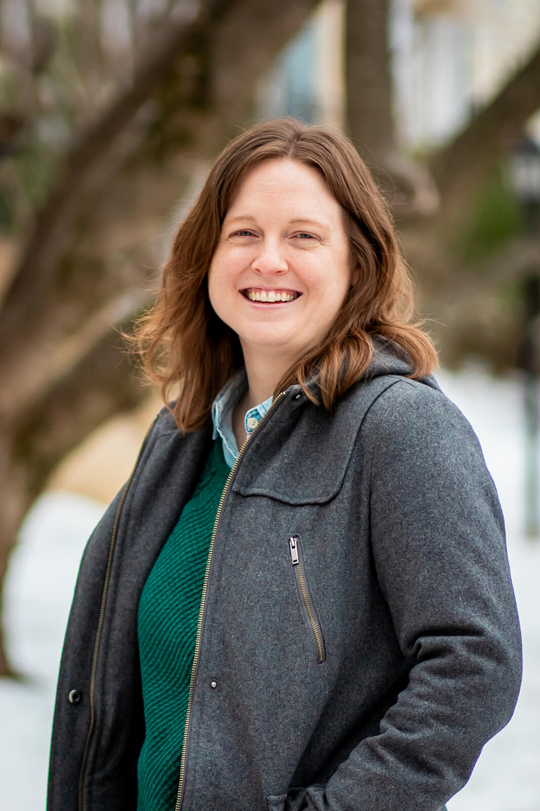 Emily Laing, LPC, a white woman in her mid to late 30s with shoulder-length auburn hair, smiles towards the camera. She wears a green sweater with a denim collared shirt underneath, jeans, and blue slippers. She sits on a blue armchair. To her right is a small side table with a lamp.