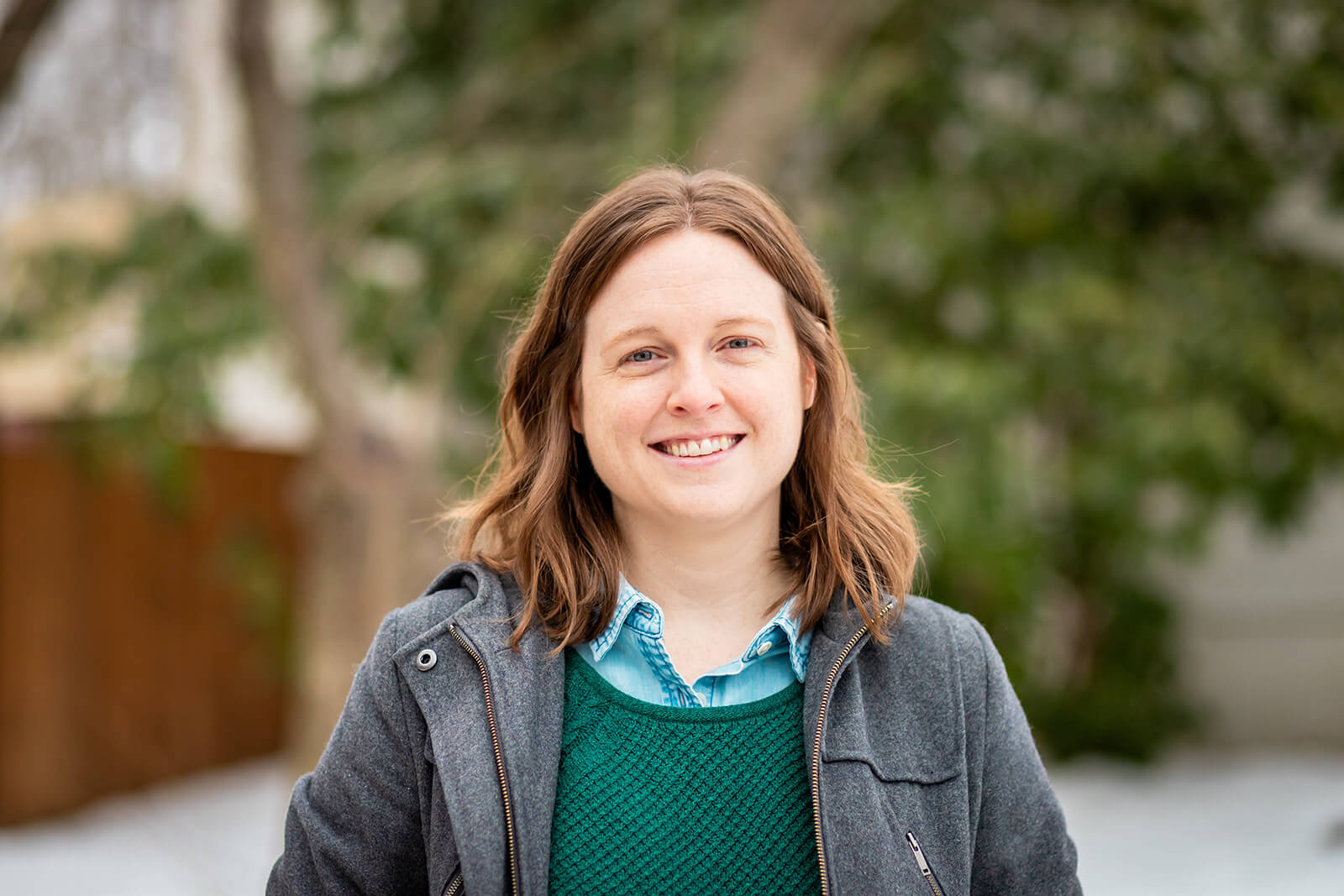 Emily Laing, LPC, a white woman in her mid to late 30s with shoulder-length auburn hair, smiles towards the camera. She wears a green sweater with a denim collared shirt underneath, jeans, and blue slippers. She sits on a blue armchair. To her right is a small side table with a lamp.