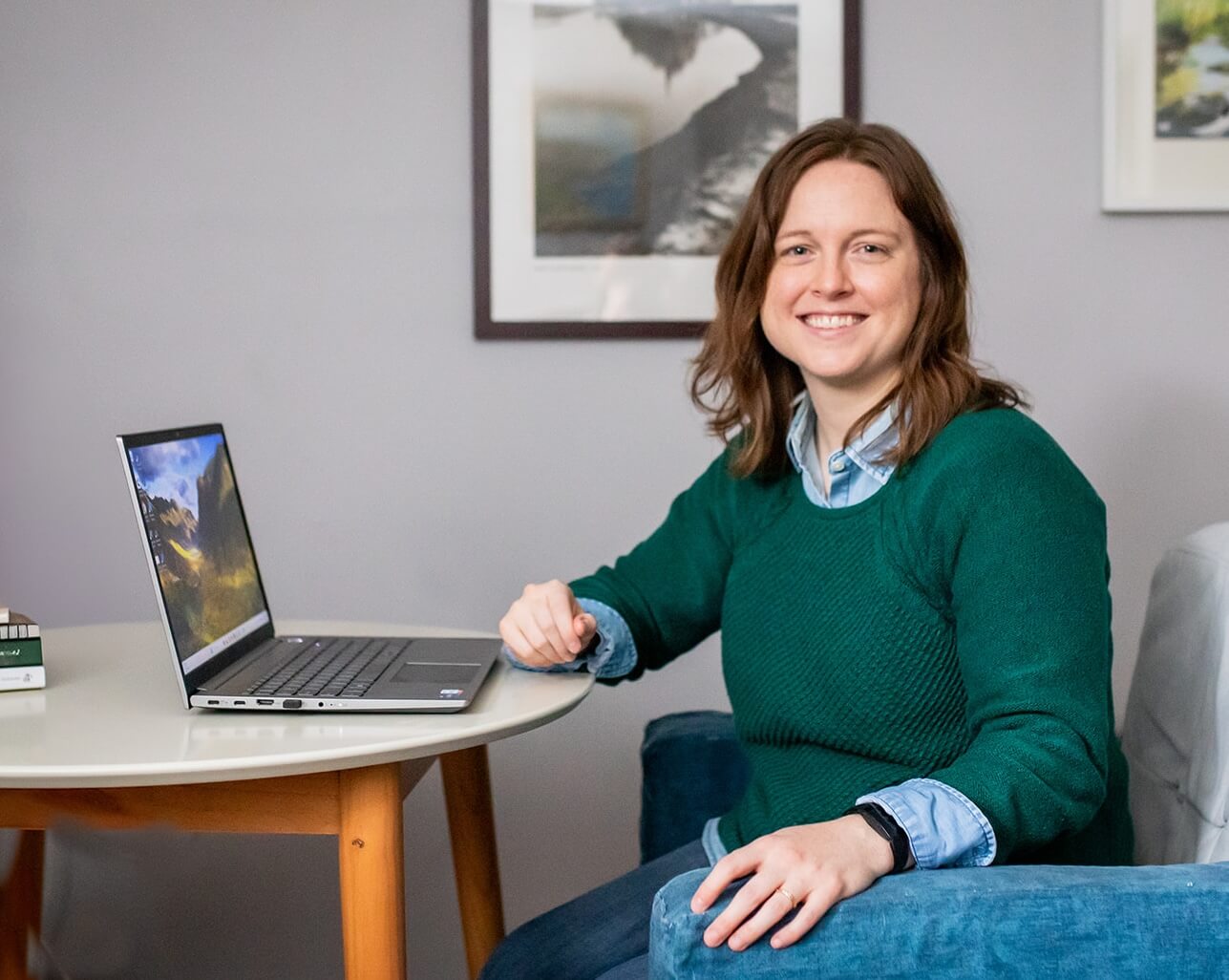 Emily Laing, LPC, a white woman in her mid to late 30s with shoulder-length auburn hair, smiles towards the camera. She wears a green sweater with a blue and white-striped collared shirt underneath. She sits at a desk with a laptop on it.