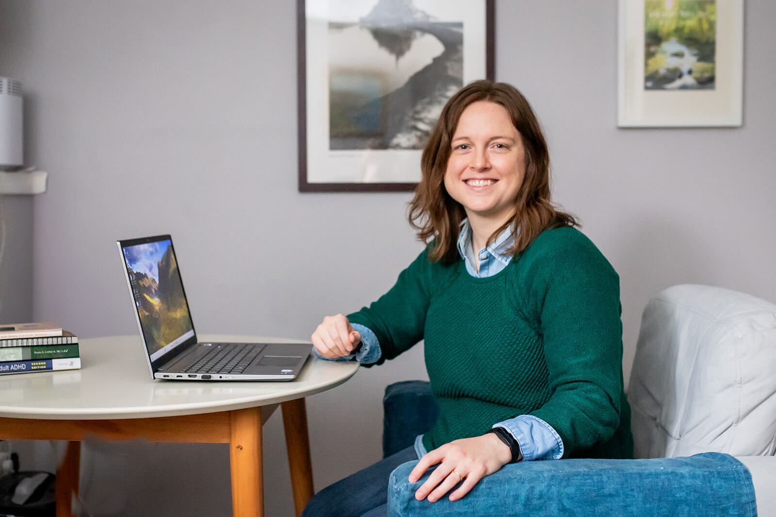 Emily Laing, LPC, a white woman in her mid to late 30s with shoulder-length auburn hair, smiles towards the camera. She wears a green sweater with a blue and white-striped collared shirt underneath. She sits at a desk with a laptop on it.